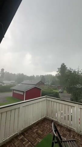 The video shows a view from a balcony overlooking a residential area during a rain shower. In the foreground, a white wooden railing surrounds a balcony with a wooden plank floor and a patch of artificial turf. A black folding chair and a round table with a small potted plant are visible on the balcony. Beyond the railing, several houses with red roofs are visible, along with trees and a street with a car parked on it. The sky is overcast and gray, indicating cloudy weather. The rain is visible as streaks falling through the air, creating a hazy effect. The overall atmosphere is calm and subdued, characteristic of a rainy day.
