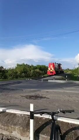 The video shows a red train crossing a railroad track at a road intersection. The train has two locomotives. The first locomotive is red with yellow accents and has the letters "РЖД" in red and white on its side. The second locomotive is also red and has a crane attached to its roof. There are two boys standing on the side of the road, watching the train. The weather is sunny with a blue sky and some clouds. The environment appears to be rural, with trees and vegetation visible in the background. There is a bicycle in the foreground of the first frame, suggesting the scene was filmed by someone on a bicycle.