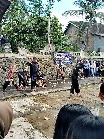 The video shows a traditional Indonesian dance performance outdoors. Several dancers, wearing colorful costumes, are performing in a muddy area. A crowd of people is watching the performance. A banner can be seen in the background with the word "RAYANDA" on it. There are houses and trees in the background, and a musical performance is happening on a porch.