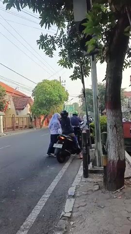 A medium shot captures a street scene with several individuals on motorcycles. A traffic light, partially obscured by tree branches, displays a red light. In the foreground, a motorcycle is parked at the side of the road. Two people, one wearing a white hijab and the other a dark top, stand beside it. Behind them, two more individuals are seated on another motorcycle. Further down the road, another motorcycle can be seen. The street is lined with buildings and trees, suggesting an urban or suburban environment. The sky is bright, indicating daylight.