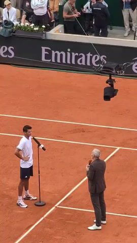 A man in a white tennis shirt and navy shorts stands with hands on his hips on a clay tennis court, speaking into a microphone. In front of him stands another man in a dark suit holding a microphone and listening. Behind them is a wall with the word “Emirates” in white lettering. Above them is a hanging camera. Around the court are spectators. The court is marked with white lines.