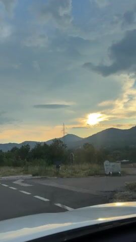 The video shows a scenic view from a car, likely a road trip. The camera is positioned from inside the car, looking out at the landscape. 

**Subjects:**
- **Scene:** The primary subject is the natural landscape, characterized by rolling hills covered in lush green trees. A road is visible in the foreground, indicating travel.
- **Objects:** A prominent electrical pylon stands tall against the sky, a sign of human infrastructure within the natural setting. There's also a white car hood in the bottom of the frame, suggesting the viewpoint.
- **Setting:** The setting is a rural or mountainous area, with a road bordering a dense forest. The hills in the background suggest a hilly or mountainous terrain.

**Activity or event:** The video captures the movement of the car, suggesting a journey or a drive through the countryside. The changing perspective of the landscape indicates motion.

**Notable details:**
- **Time of day:** The presence of the sun low on the horizon, casting a warm glow and creating long shadows, suggests it is either sunrise or sunset. The sky is partly cloudy, with dramatic cloud formations.
- **Weather:** The sky appears to be partly cloudy, with the sun breaking through. The light suggests a pleasant, possibly warm day.
- **Location cues:** A small signpost with white and red markings is visible next to the road, typical for road markers. While no specific text is clearly legible, the overall environment is consistent with a rural or mountainous region.
- **Emotions:** The scenic view evokes a sense of peace, tranquility, and the freedom associated with travel. The golden light from the sun adds a touch of warmth and beauty.

**Visible text:** There is a small signpost by the road with some text, but it is not legible enough to be read. There is also some text on the pylon, but it is too small to be deciphered.