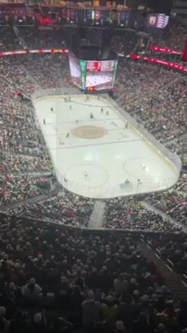 The video shows a wide, high-angle view of an ice hockey game in a large, packed arena. The rink is centrally located, with players skating and interacting on the ice. The crowd fills the surrounding seats, mostly wearing casual clothing. A large screen hangs above the center of the rink, displaying the game's score and possibly replays. Banners and flags are visible in the upper areas of the arena. The lighting is bright, illuminating the ice and the audience.