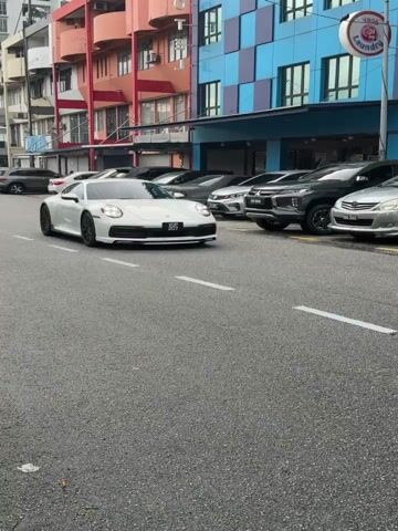 The video shows a white Porsche driving down a street in a city. The Porsche is traveling from right to left in the frame. The car has black wheels and a black front lip spoiler. There are other cars parked on the side of the street. The buildings are a variety of colors and styles, including blue, orange, and white. There is a laundry business, and one building has a movie theatre on its side.  The road has a single dashed white line separating the lanes of traffic.