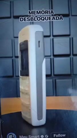 The media file shows a close-up, slightly blurry, side-view shot of a white Nokia 1100 mobile phone. The phone is positioned on a dark, textured surface, possibly a table or a platform, with a hint of an orange surface beneath it. In the background, there's a dark, grid-like pattern, suggestive of a textured wall or screen.

The phone's screen is illuminated with a bright blue display, showing text in Portuguese. The text at the top reads "MEMÓRIA DESBLOQUEADA," which translates to "MEMORY UNLOCKED." Below this, the screen displays a list of items, possibly song titles or menu options. Some of the visible text includes "Toque Musical," "Espionage," "Everlasting," and "Liszt." There are also options like "OK" and "Voltar" (Back).

The phone itself appears to be a classic model, with a visible keypad and navigation buttons. The casing shows some signs of wear, with a slightly yellowed or discolored appearance in certain areas. The overall lighting suggests an indoor setting, with the primary light source illuminating the phone and its screen. The visual quality of the video is not high-definition, with some artifacts and a slightly shaky camera movement.

There are no people visible in the frame, and the focus is solely on the mobile phone and its display. The context of "Mubi, Nigeria" is external information and not visually evident within the media file itself. The video seems to be a demonstration or a showcase of the phone's functionality or its unlocked memory status.
