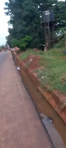 The video shows a street scene from a slightly low angle. The road is a reddish-brown color and has a concrete ditch running alongside it. Water is present in the ditch. Grass and red dirt are present on the embankment between the ditch and the road. In the background, a water tower stands among trees. As the video continues, the camera pans to the left to reveal people in the distance walking and working along the road. The sky is overcast.