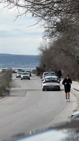 The video captures a street scene in an outdoor setting. A man dressed in black shorts, a black hoodie, and white socks is jogging along the right side of a paved road, wearing what looks like headphones. Cars are parked and driving on the left side of the road towards the viewer. The landscape includes a body of water with visible mountains in the background, and bare trees line the side of the road. Other pedestrians are seen walking near the parked cars. The sky is overcast, indicating it may be a cloudy or overcast day.