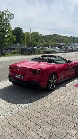 The video shows a red Ferrari Portofino convertible pulling away from the curb, onto the road and driving into traffic. A man in a dark vest walks from right to left along the sidewalk as the car pulls away. The Ferrari has a license plate that reads "WT Q 63." The road is lined with parked cars, a building, trees, street signs, and traffic lights.  The sidewalk is made of light-colored bricks. A red carpet is partially visible. The sky is bright with clouds.