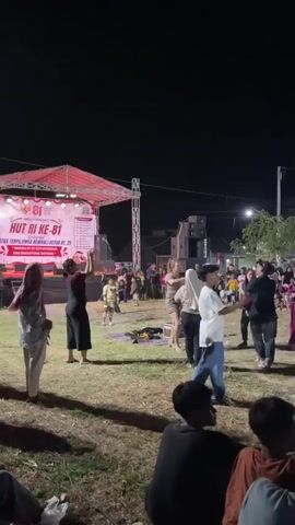 The video shows an outdoor nighttime event with a stage set up on the left. The stage has red lighting and a banner that reads "HUT RI KE-81" which translates to "81st Anniversary of the Republic of Indonesia." Several people are gathered in front of the stage, with some individuals on the stage performing. In the foreground, the backs of several people are visible, appearing to be spectators sitting or standing. The environment appears to be a grassy field with trees and buildings in the background. The lighting is artificial, primarily from the stage and surrounding lights. The atmosphere seems festive, with people interacting and watching the performance.