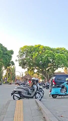 The video begins by showing a street scene with several parked motorcycles. A large tree with lush green foliage is visible on the right side of the screen. As the video progresses, the camera pans to the left, revealing a traditional Balinese temple-like structure with intricate architecture. People are seen sitting and standing in front of the temple, and more motorcycles are parked along the roadside. A few people are riding motorcycles on the street. The sky is clear and bright, suggesting it is daytime. The overall scene depicts a public space with cultural elements, traffic, and people engaged in various activities.