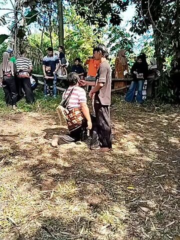 The video shows two men in a clearing surrounded by trees, with a crowd of people watching from the edges. One man wears a patterned shirt and is initially on his knees, then stands up. The other man wears a dark shirt and a patterned skirt. They appear to be engaging in a performance or ritualistic interaction. The ground is covered with dry leaves and grass. In the clearing there is also a black container on the ground. The crowd includes people of various ages and clothing styles. The atmosphere suggests a cultural event taking place outdoors in a rural area.