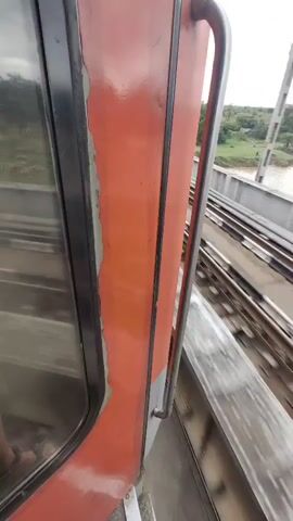 A shaky, first-person perspective video shows a red and gray train car with a metal handrail. The train appears to be in motion, with the tracks and surrounding landscape blurred in the background. The exterior of the train shows signs of wear and tear, with peeling paint on the red section. The environment outside the train consists of green vegetation, a body of water, and a bridge structure. The video is short, lasting only a few seconds.