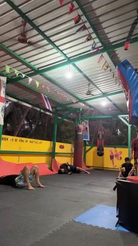 The video shows an open-air gym space with a tin roof and green structural supports adorned with international flags. Multiple people are scattered across the black floor mats, some sitting and stretching. Several punching bags hang in the background.  A woman is seen lying on the mat, wearing a pink sports bra and lavender shorts, holding the camera. She appears to be taking a selfie or video of herself. The gym seems to be lightly populated, with people engaged in individual activities.