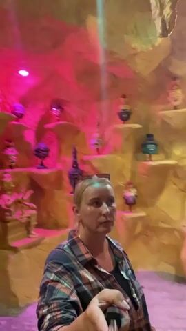 A woman with blonde hair, wearing a plaid shirt and a black tank top, is holding a selfie stick and looking directly at the camera. She is in a cave-like environment with rough, textured walls that are illuminated with pink and yellow lights. The walls are adorned with shelves displaying various objects, including what appear to be statues, masks, and decorative vases. Some of these items are golden or have gold accents. In the background, to the left, there is an arched opening leading to another area where another person is visible, walking away from the camera. The lighting is dramatic, with spotlights creating beams of light that fall on the cave walls and objects. The overall impression is that of a decorative, perhaps tourist-oriented, cave or grotto.