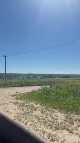 The video shows a view from inside a vehicle looking out at a rural landscape on a sunny day. The sky is clear blue with a bright sun. In the foreground, there is a dirt road and the edge of the vehicle's interior, possibly a window frame, is visible on the left. Beyond the road is a field of green grass with some taller weeds and small plants. A single utility pole with power lines stretches across the frame, slightly off-center to the left. In the distance, there is a line of trees and buildings, suggesting a small town or village. The sun is positioned high in the sky, casting a bright light and creating a slight lens flare effect. The overall impression is a serene, open, and sunny rural environment.