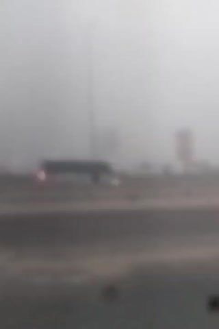 The video begins with a view of a road obscured by thick fog. A bus is visible in the distance, its headlights shining through the mist. The camera pans slightly to the right, revealing more of the road and the foggy environment. Signs and streetlights are faintly visible through the fog. The video continues to show the road stretching into the distance, with the fog consistently limiting visibility. Structures and possibly more vehicles can be seen dimly through the haze on the right side of the road. The scene remains consistent throughout the video, characterized by heavy fog and low visibility on a road.