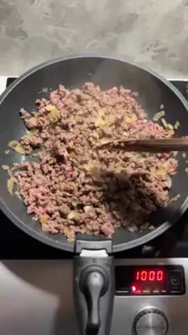 A person is cooking ground beef and onions in a black non-stick frying pan on an electric stove top. The ground beef is mostly cooked and brown with some pink still visible. The onions are translucent and mixed in with the beef. The person is using a wooden spatula to stir the mixture in the pan. The stovetop setting is set to 1000.  The background is a gray countertop.