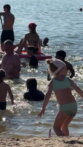 The video captures a sunny day at a lake, likely in Malakhovka, Russia, with numerous people enjoying recreational activities. The scene is dominated by the sparkling water, reflecting the bright sunlight, suggesting a warm, clear day, possibly late morning or early afternoon given the intensity of the light.

The primary activity is swimming and wading in the lake. A young girl in a light green bikini is prominently featured in the foreground, facing away from the camera and towards the water. She appears to be walking or standing at the water's edge. In the background, various individuals are visible. Some are swimming or submerged in the water, while others are interacting on paddleboards. A woman with a red bandana is seated on a paddleboard, and another person is paddling a board with an oar. A man in the mid-ground is standing in the water, seemingly interacting with others. Further back, several people are in the water, some appearing to be children and adults. There's also a glimpse of what appears to be a dark-colored dog swimming in the water.

The mood seems relaxed and joyful, typical of a summer outing at a lake. The sunlit water and the presence of multiple groups of people suggest a communal and leisurely atmosphere. There are no visible signs or text within the video clip. The overall setting is a natural body of water with a sandy or dirt shoreline in the immediate foreground.