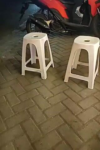 The video captures a street scene at night. In the foreground, two white plastic stools are placed on a brick-patterned walkway. Behind the stools, a red motorcycle is parked. The background shows a street with some shops and a mosque with illuminated minarets. Vehicles, including a truck and a motorcycle, are visible on the street. The lighting suggests it is nighttime, with artificial lights from the shops, mosque, and vehicles illuminating the scene.