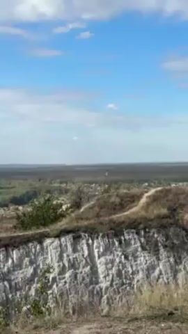 The video shows a wide-angle view of a landscape under a blue sky with scattered white clouds. In the foreground, there is a steep, light-colored cliff face with textured, layered rock formations. The top of the cliff is covered with dry grass, sparse vegetation, and a dirt path. The background consists of a dense forest stretching to the horizon, with occasional buildings or settlements visible amidst the trees. In the far distance, there appears to be a faint white structure, possibly a factory or a large building. A single yellow and green flag is visible on the horizon.