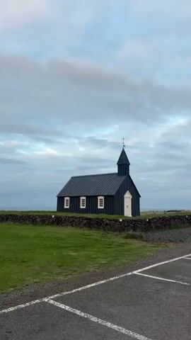 The media features a wide shot of the iconic black church of Búðir, Iceland, positioned centrally within the frame.

**Subjects:** The main subject is the small, dark-colored wooden church, characterized by its pointed steeple topped with a cross, a dark corrugated roof, and a striking white main door. Three white-framed windows are visible on the church's left side. The church stands on a vibrant green grassy patch, which is enclosed by a low, dark stone wall.

**Scene and Setting:** The scene is set outdoors under an expansive, cloudy sky. Beyond the church and the stone wall, the horizon line of what appears to be the ocean is faintly visible. In the foreground, a section of an asphalt parking lot with white painted lines indicating parking spaces occupies the lower portion of the frame.

**Activity or Event:** There is no discernible activity or event taking place; the scene is static, captured with only a very subtle, almost imperceptible camera movement.

**Notable Details:** The sky is overcast, providing soft, diffused light that creates a serene and somewhat muted atmosphere. The time of day is not clearly indicated but the gentle light suggests either early morning or late evening rather than harsh midday sun. The distinct architecture of the church and its isolated coastal setting contribute to a sense of peaceful solitude and stark beauty.

**Visible Text:** There is no visible text present in the video.