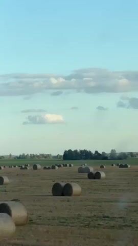 A drone footage captures a rural landscape with a harvested field. Numerous round hay bales are scattered across the field. In the distance, a line of trees forms a horizon, and the sky is a pale blue with scattered clouds. The overall lighting suggests it is daytime.