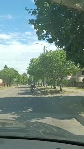 This video captures a sunny daytime scene from the perspective of a vehicle traveling down a tree-lined suburban street. The sky is bright blue with scattered white clouds. The road is asphalt, with a clear lane marking visible. On the right side of the road, there is a sidewalk and grassy areas bordered by trees and a fence, suggesting residential properties. On the left, more trees and some buildings are visible in the distance.

The primary activity is a person riding a white scooter down the middle of the road, moving away from the camera. The rider appears to be wearing a light-colored top and dark bottoms, with a helmet. The scooter is a typical model, with a passenger seat. The overall impression is a casual, everyday moment of travel on a pleasant day.

The time of day is clearly daytime, indicated by the bright sunlight and the clear visibility. The weather appears to be warm and fair, conducive to outdoor activities like riding a scooter. The setting is suburban, characterized by the presence of trees, houses, and residential infrastructure like streetlights and power lines. The lack of significant traffic suggests a relatively quiet area. There is no visible text in the media file. The mood is tranquil and ordinary, depicting a simple slice of life.