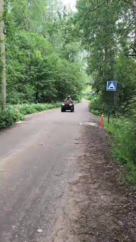 A quad bike with two people on it drives down a paved road. The road is lined with trees on both sides, and there is a white sign with a blue triangle on the right side of the road. The quad bike is red and black, and the people are wearing helmets and camouflage clothing. The weather appears to be overcast.