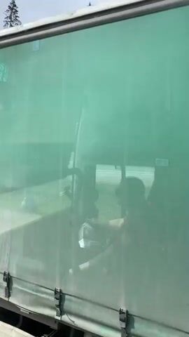 The video shows a green semi-truck with white lettering. Initially, the side of the trailer is visible, which reads "ORTE CORP". The camera then pans to the front of the cab, revealing a sign that says "ПРИГЛАШАЕМ ВОДИТЕЛЕЙ ИЗ РЕГИОНОВ РФ 8 800 700 84 48". The camera continues to move forward, capturing the view from the passenger side of a vehicle driving down a road. The road is marked with white lines and arrow symbols. A bus stop and a gray metal sound barrier are visible on the side of the road, along with a forested area in the background.