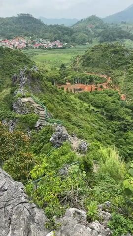 A drone shot pans across a lush green mountainside. In the foreground, large gray rocks are covered in thick green vegetation. A stone staircase with a bright green metal railing winds its way up the rocky incline. Beyond the staircase, more green hills stretch into the distance, dotted with trees. In the far background, a village with red-tiled roofs is nestled in a valley. The sky is overcast.