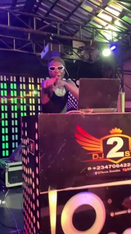 A young black male DJ stands behind a DJ console at what appears to be a club or outdoor venue. He wears a black tank top and futuristic white-rimmed sunglasses. The DJ's console is black and decorated with a logo featuring a set of wings and the letters "DJ" above a number, along with a phone number and social media handle. A laptop sits on the console in front of him. Behind the DJ is a wall with a colorful, rectangular light display, and above him are various lighting fixtures, including a disco ball. The lighting shifts in color, illuminating the DJ in shades of pink, blue, and green. The DJ is actively performing, moving his hands in coordination with the music and appearing to be dancing.