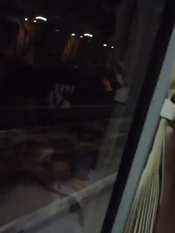 The video is shot from inside a vehicle, looking out a window at night. The scene outside is dark, with some lights visible in the distance, possibly streetlights or lights from buildings. A curtain is visible on the right side of the frame. The window shows a road with yellow lane markings. There appears to be a sign with a red circle and a white 'P' on it, indicating parking. The overall impression is of a journey through a dimly lit urban or suburban area at night.