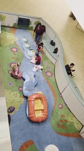 The video captures an overhead view of a children's play area, likely in a mall or similar indoor public space. The floor is a colorful carpet depicting a whimsical river scene with flowers and trees. In the center of the play area, a blue, wavy slide structure is present, with a red raft-like toy at its base. There are several people in the scene. In the upper right, a person in a dark maroon shirt and blue jeans stands, appearing to observe or guide. To their left, a person in a black shirt and jeans sits on a curved bench. Further down the bench, another person in a black shirt and white pants sits, looking at a phone. On the carpeted play area, a person in a red and white top and dark shorts is seated near the top of the slide, with a baby or small child in white lying in front of them. To their left, a person in a pink and white patterned dress is kneeling, looking towards the slide. To the right of the seated person in red, a child in a red shirt and dark pants is crawling or walking. The overall impression is a relaxed, everyday scene in a family-friendly environment.
