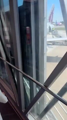 A young boy wearing a Spider-Man backpack walks through an airport jet bridge, followed by a woman. The jet bridge is enclosed with glass on one side, offering a view of the tarmac. Other passengers and airport staff are visible in the background.