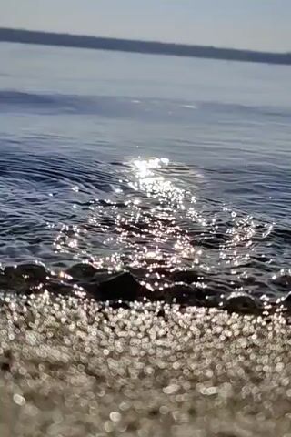 The video shows a close-up view of a body of water, likely a lake or ocean, with the sun reflecting on its surface. The foreground is a sandy or pebbly shore. The water is calm with gentle ripples. The sun's reflection creates a shimmering, bright path across the water. There are no people or animals visible in the video. The environment appears to be natural and serene.