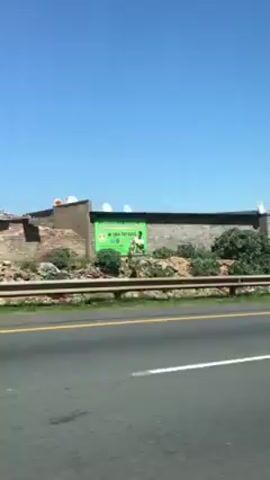 The video shows a road scene, likely filmed from inside a moving car. The sky is clear blue. The roadside is visible with a metal guardrail. Beyond the guardrail, there is a small slope and vegetation with some rudimentary buildings or shacks constructed from what appear to be cinder blocks. Some structures have satellite dishes.  The landscape looks dry and somewhat barren. A large white frame structure passes overhead on the road.