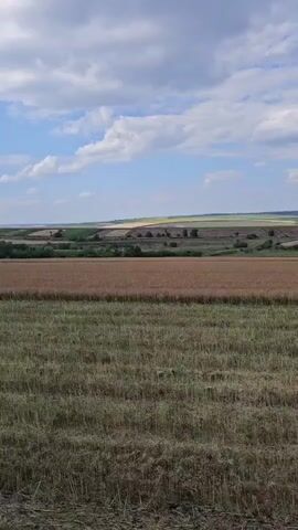 A tractor is driving through a field. The field is mostly dry grass and stubble. In the background there are hills and more fields. The sky is blue with some clouds.