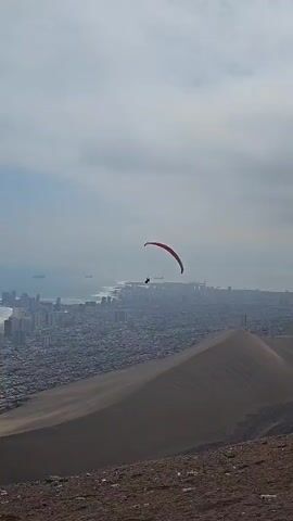 A paraglider flies over a sand dune. The paraglider has a red canopy and the pilot is silhouetted against the sky. Behind the dune is a large city on the coast, obscured by haze. Several ships are visible on the water in the distance. The foreground shows the top of a dune with rocky, barren ground. The sky is overcast.