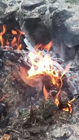 The video shows a close-up shot of a small fire burning amongst rocks and debris. Flames are visible, ranging in color from yellow to orange, and some smoke is rising. The materials burning appear to be wood, paper, and possibly some plastic. The overall scene is dark, with the fire providing the primary source of light.