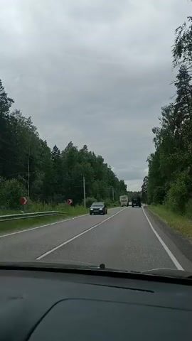 The video shows a view from inside a car driving on a paved road. The road is lined with trees and appears to be in a rural or wooded area. There are other vehicles on the road, including a dark-colored sedan on the left side of the road, a white truck with a covered cargo bed, and a dark-colored SUV or truck further ahead. The sky is overcast. The car is moving forward at a moderate pace.