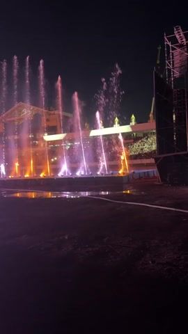 The video shows an outdoor nighttime scene with a stage and a crowd of people in the background. In the foreground, there are several water fountains spraying water upwards, illuminated with pink and orange lights. The fountains are arranged in a line, with some smaller fountains in front and taller ones behind. Behind the fountains, there is a large stage structure with lights and what appears to be a large audience seated or standing. The overall atmosphere suggests a concert or a public event with a light and water show. The ground in the foreground is dark and appears to be dirt or grass, with some puddles of water reflecting the lights. To the right, a large metal structure, possibly a scaffolding or lighting rig, is visible. The sky is dark, indicating nighttime.
