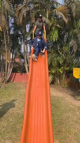 Two boys are playing on an orange slide in a grassy park. One boy, wearing a blue denim jacket and a baseball cap, slides down, landing face-first on the ground. The other boy, wearing a green shirt and a baseball cap, waits at the top and then slides down headfirst.  Palm trees and other greenery surround the play area.  A red brick wall is visible in the background.