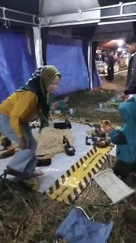 A person wearing a yellow hoodie and blue jeans is seen bending down and interacting with objects laid out on a white tarp. They are wearing a hijab. A child in a blue hooded jacket is standing to the right and looking at the objects on the tarp. Behind them, two individuals, one in a black jacket and the other in white pants, are visible. The scene appears to be outdoors, possibly under a tent, with blue tarps in the background and dry grass on the ground. There are various objects on the tarp, including small toy vehicles and what appears to be grain or seeds. A black and yellow caution tape is also present on the tarp.