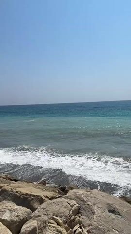 The video shows a rocky coastline with the ocean and a clear blue sky. Waves are crashing on the shore. The water is a gradient of colors, from a light turquoise near the shore to a deep blue further out. In the distance, a coastline with buildings can be seen. The video appears to be taken during the daytime on a sunny day. There are no people or animals visible in the video.