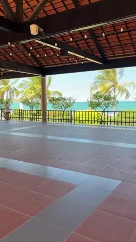 The video pans across a tiled patio area with a view of palm trees, lush green bushes, a black metal railing, and a bright blue ocean under a clear blue sky. A person in a light-colored shirt and dark shorts stands on the far left of the patio, looking out at the ocean. String lights are visible along the underside of the covered patio ceiling.