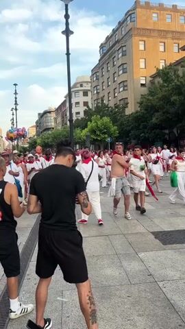 The video captures a lively street scene in a European city, likely during a festival or celebration. A large crowd of people, predominantly dressed in white clothing with red accents like bandanas and sashes, fills the cobblestone street and sidewalks. The atmosphere appears festive, with many individuals smiling and interacting. In the foreground, a person with dark hair tied in a bun and tattoos on their arm is looking at their phone. To their left, another person in a black tank top with the number 13 on it stands. Further back, the crowd stretches along the street, flanked by buildings with balconies and windows. Some people are holding balloons, and others are carrying objects that resemble fans. The sky is partly cloudy, and the sun is casting a warm light, suggesting it's either morning or late afternoon. The overall impression is one of a vibrant, communal event.
