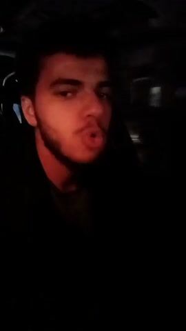 A young man with dark hair and a trimmed beard is visible from the chest up, inside a car at night. The light inside the car is dark and intermittently turns red, presumably from the tail lights of other vehicles. The man is looking to the right and moving his lips, as if speaking or making noises. The background outside the car window is blurry and indistinct due to the darkness and motion, but suggests he is driving on a road in a city.