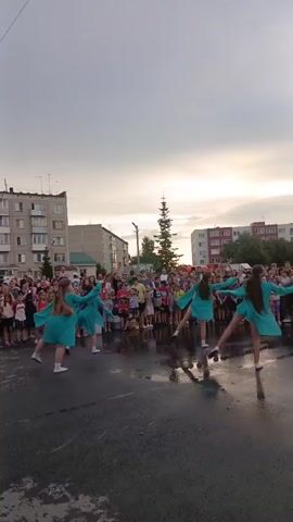 A group of young girls in matching teal dresses are performing a synchronized dance routine on a wet asphalt surface in front of a crowd of onlookers. The dancers, dressed in loose, knee-length tunics and white socks, move in unison with flowing arm gestures and coordinated steps. The background features apartment buildings and a tall evergreen tree, suggesting an outdoor urban or town setting. The sky above is overcast with hints of light breaking through. The crowd, composed of adults and children, watches the performance attentively. The ground is wet with puddles, reflecting the sky and the dancers. The overall atmosphere appears to be that of a community event or celebration.