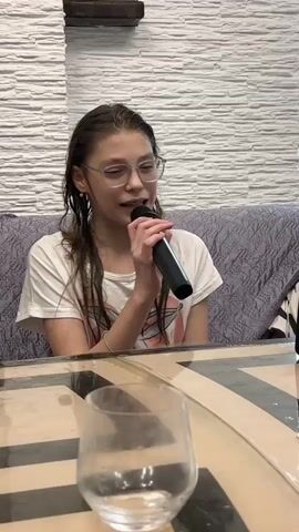 A young person with wet hair and glasses sings into a microphone, sitting at a table with a glass on it. The background includes a textured white wall and a patterned sofa.