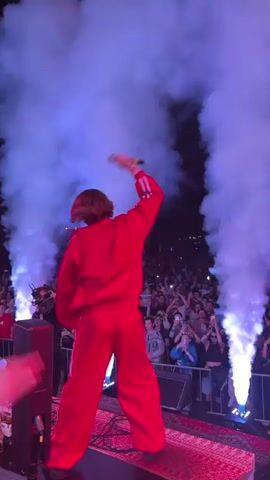 A person in a red jumpsuit with white stripes stands on a stage, facing away from the camera and towards a large crowd. They have their right arm raised with a fist in the air. There are plumes of white smoke on either side of the person, illuminated by blue and pink lights. The crowd is packed behind a barrier, with many people holding up their phones to record. The background is dark, suggesting it is nighttime.